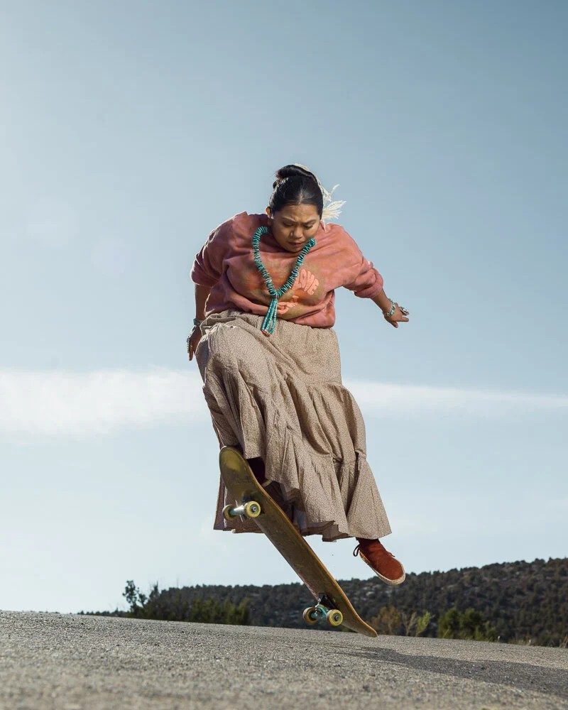 A woman in a traditional skirt and turquoise jewelry skateboarding mid-air on a desert road, photographed by Tomás Karmelo Amaya